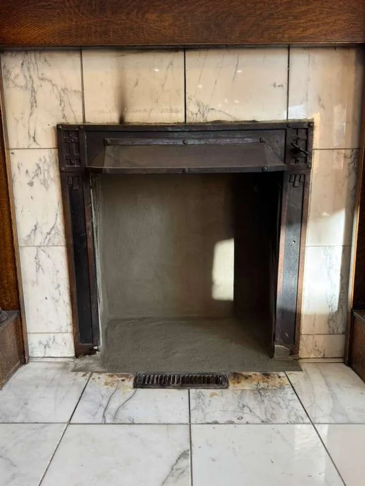 A white marble fireplace with a dark iron frame, an oak mantel above, a clean empty firebox and a small floor vent in the hearth