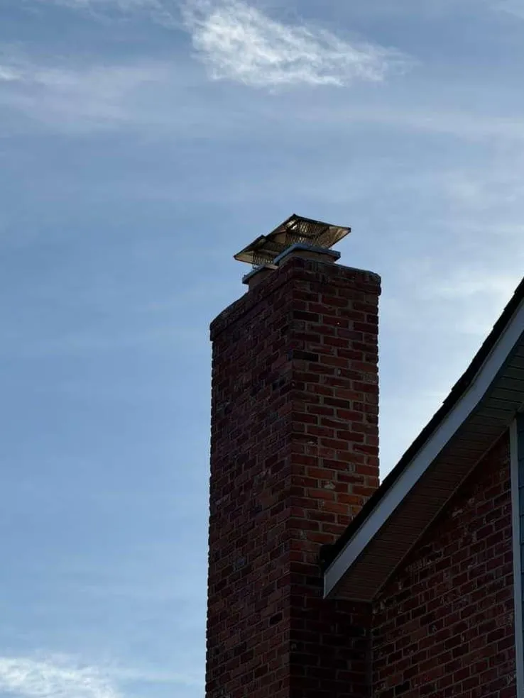 A tall red brick chimney against a blue sky, topped with a shiny steel cap, beside the white eave of a house with blue siding