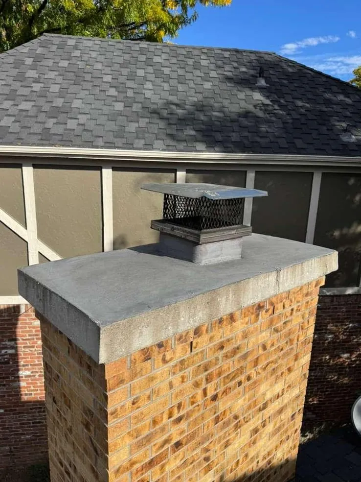 A tan brick chimney with a gray concrete crown and a black mesh cap, beside a house with a dark shingle roof