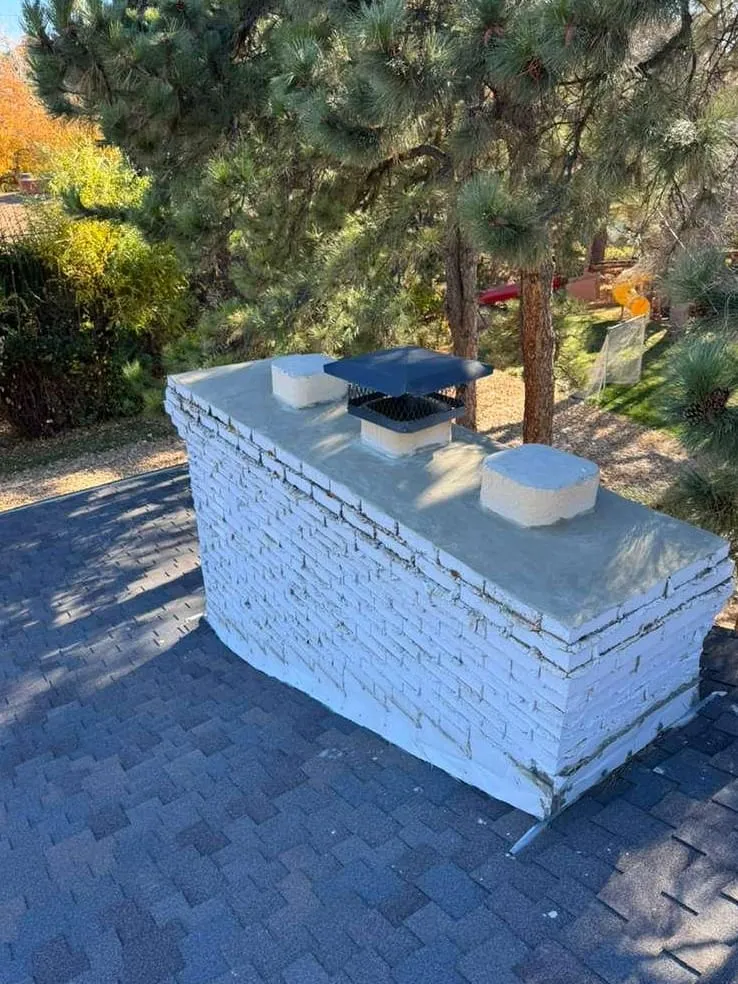 A chimney of white-painted brick with two round clay flue tops and a flat blue cap on a concrete crown, with pine trees behind and a gray shingle roof around it